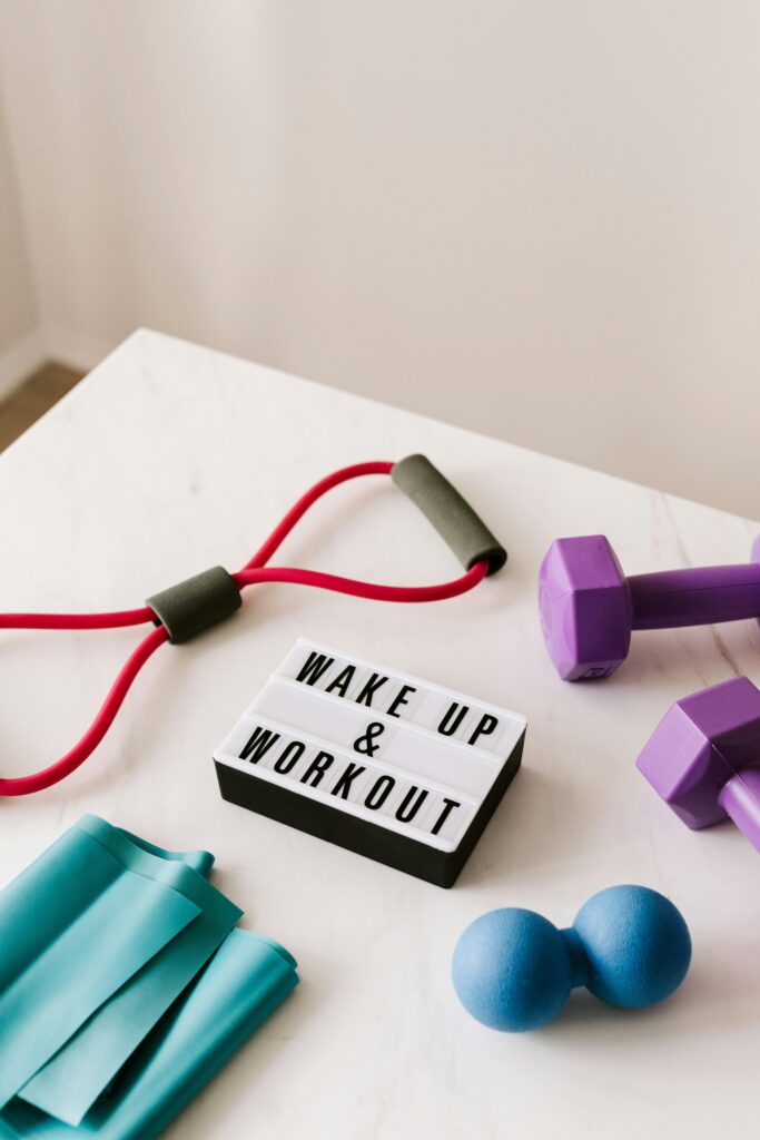 From above composition of dumbbells and massage double ball and tape and tubular expanders surrounding light box with wake up and workout words placed on white surface of table.Best online women's fitness training kerala